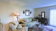 This single-floor space showcases a thoughtfully decorated living room that combines classic elegance with a warm, comfortable ambiance. The room is anchored by a sage green sofa adorned with patterned throw pillows, complemented by floral-upholstered accent chairs that add a layered, traditional aesthetic. Neutral-toned walls and smooth ceilings provide a clean backdrop that allows the decorative elements to stand out.

Ornate wrought-iron wall sconces flank a framed mirror, adding a touch of Old World charm to the space. A sculptural table lamp with a fabric shade contributes to the warm lighting scheme. The glass-topped coffee table with an intricate wrought-iron base serves as a focal point, reinforcing the room's refined style.

A dark wood console table along the back wall is styled with decorative objects and greenery, adding depth and visual interest. A large mirror above it further enhances the sense of space. The flooring features warm terracotta-toned tiles, grounding the room with a Mediterranean-inspired character. Overall, the space presents a cohesive, traditionally styled interior with attention to decorative detail.