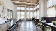Located at 180 Woodruff Rd in Greenville, SC, this single-floor community living room is a well-appointed multipurpose lounge space designed to serve both work and leisure needs. The room immediately impresses with its dramatically high ceilings, which are accented by warm wood beam detailing and pendant lighting that adds a refined, modern touch overhead.

Expansive floor-to-ceiling windows line the walls, dressed with layered sheer and woven curtains that filter abundant natural light while maintaining a soft, welcoming ambiance. The transom windows above further amplify the sense of openness and height throughout the space.

Rich wood-look plank flooring runs throughout, grounding the room with a warm, contemporary aesthetic. Along one side, a built-in workstation with desks, computers, and ergonomic seating provides a dedicated area for productivity. On the opposite end, a dark upholstered sofa anchors a casual lounge zone, complemented by round tables and plush green accent chairs that invite conversation and relaxation.

Glass-paneled dark-framed doors provide access to adjacent areas, contributing to the open, airy feel of the space. A wall-mounted television and a media console further enhance the room's functionality. Overall, this living room offers a polished, comfortable, and practical environment suited to a variety of community activities.