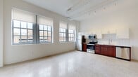 This single-floor unit showcases a distinctive industrial-loft aesthetic, blending modern finishes with architectural character. The open-concept living and kitchen area is anchored by polished terrazzo flooring that extends throughout, while exposed cylindrical ductwork and track lighting run across the elevated ceiling, reinforcing the space's urban character. A generous bank of large, grid-paned windows lines the main living area, flooding the space with natural light and offering elevated views of the surrounding cityscape.

The kitchen is thoughtfully designed with rich cherry-toned lower cabinetry paired with crisp white upper cabinets, a classic white subway tile backsplash, light countertops, and a full suite of stainless steel appliances including a refrigerator, range, microwave, and dishwasher. The entry door features a glass panel, adding additional light near the kitchen.

The bedroom is a generously sized, carpeted room with tall ceilings and large windows that continue the urban view. A spacious walk-in closet provides ample storage with full-length hanging rods. The bathroom features a dual-sink vanity with white countertops, a large mirror, and conveniently integrated in-unit stacked washer and dryer, all finished with the same terrazzo flooring found throughout the unit.