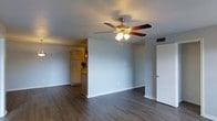 This single-floor space offers a straightforward, functional layout centered around an open living area that flows visually toward the kitchen. The living room features dark hardwood-style flooring that contrasts cleanly with the light gray walls and white baseboards, creating a neutral, modern aesthetic. A ceiling fan with an integrated light fixture is centrally mounted, providing both air circulation and ambient lighting to the space.

The walls are smooth and painted in a soft gray tone, lending the interior a calm, understated character. A standard entry door is positioned along one wall, offering easy access to the unit. Overhead, a ceiling-mounted air vent indicates a central HVAC system is in place.

Partially visible from the living area, the kitchen features light wood cabinetry and appears to include a full-size refrigerator. The open sightline between the two spaces enhances the sense of connectivity throughout the unit. The overall design is practical and clean, making it a functional space suitable for everyday living.