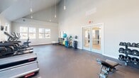 Located at 35 Mussey Rd in Scarborough, Maine, this fitness center offers a bright, modern workout environment designed to support a wide range of exercise routines. The space is defined by its dramatic vaulted ceiling, which rises steeply to a peak and lends an open, airy quality throughout. A bank of large, multi-pane windows along the main wall floods the interior with natural light and provides views of the surrounding area, creating an energizing atmosphere for workouts.

The flooring consists of durable, dark rubberized material suited for high-impact activity, while the neutral gray walls and clean white trim give the space a polished, contemporary look. Pendant lighting fixtures hang from the vaulted ceiling, complementing the natural light and ensuring the space is well-lit at all times.

The gym is outfitted with multiple treadmills and elliptical machines arranged along the window wall, along with a multi-station cable strength machine and an adjustable weight bench. A dedicated area near the entry features open shelving, a seating bench, and a well-stocked rack of exercise accessories including stability balls and resistance equipment. Glass double doors provide access to an adjacent area, contributing to an open and connected layout. Overall, the space delivers a fully functional and inviting fitness experience.