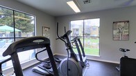 This single-floor fitness room offers a dedicated space for cardiovascular exercise, designed with both functionality and comfort in mind. The room is outfitted with key cardio equipment, including a treadmill and an elliptical machine, catering to a range of workout preferences. Large windows line the walls, flooding the space with natural light and offering pleasant views of the well-maintained outdoor grounds, complete with green lawn areas, a small pond, and mature trees. The bright, airy atmosphere is further supported by overhead fluorescent lighting, ensuring the space remains well-lit at all times. The walls are finished in a clean, neutral tone, contributing to a focused and motivating workout environment. Dark carpeted flooring provides a practical surface underfoot, helping to absorb impact and reduce noise. A fitness reference poster displayed on the wall adds a functional touch to the interior. The generous window placement not only enhances the ambiance but also keeps the space feeling open and connected to the outdoors, making workouts a more enjoyable experience.