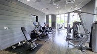 This fitness center offers a thoughtfully designed workout environment spread across a single level. The space features hardwood-style flooring and a drop ceiling with recessed lighting and ceiling fans, ensuring comfort during exercise. One wall is finished with horizontal shiplap-style paneling in a neutral grey tone, adding a modern, clean aesthetic to the interior.

The facility is well-stocked with a variety of cardio and strength-training equipment, including ellipticals, stationary bikes, cable machines, and adjustable benches, catering to a wide range of fitness routines. Equipment is arranged to allow ample circulation space between machines.

Large floor-to-ceiling windows along one wall bathe the room in natural light and provide open views of the surrounding greenery and skyline, creating an energizing atmosphere. The combination of modern finishes, abundant natural light, and comprehensive equipment selection makes this fitness center a functional and inviting space for residents or members looking to maintain an active lifestyle.