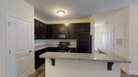 This single-story, two-bedroom, two-bathroom residence offers a thoughtfully designed layout that balances functionality and comfort throughout. The kitchen serves as a central highlight, featuring rich dark cabinetry with sleek hardware, light granite countertops, and a full suite of black appliances including a range, microwave, and refrigerator. A peninsula with an undermount double sink and breakfast bar overhang connects the kitchen to the main living area, making it ideal for casual dining or entertaining. Wood-style plank flooring runs through the kitchen, living area, and bathrooms, lending a cohesive and contemporary feel. The living room is bright and open, with natural light from a window, a ceiling fan with lighting, and ample space for varied furniture arrangements. Both bedrooms are carpeted for warmth and comfort, each featuring ceiling fans, natural light from windows, and generous floor space. The two bathrooms mirror each other in style, each offering dark vanity cabinetry, granite countertops, undermount sinks, and wood-style flooring. One bathroom includes a tub-shower combination. Overall, the unit presents a clean, modern aesthetic with consistent finishes throughout.