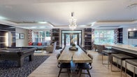 This thoughtfully designed single-floor amenity space combines multiple functional zones within an open, cohesive layout. The main gathering area serves as the centerpiece, anchored by a striking live-edge communal table beneath a modern chandelier, surrounded by upholstered dining chairs. Tray ceilings with recessed lighting add architectural depth throughout, while light-toned hardwood-style flooring unifies the space.

A lounge zone features a teal sofa accented with bold red and patterned throw pillows, flanked by sleek dark built-in shelving units and a flat-screen television. Large windows dressed in graphic red-and-white curtains flood the area with natural light and offer views of a landscaped outdoor courtyard. Glass doors provide direct access to the exterior.

The game area accommodates a full-size billiards table positioned atop a navy patterned area rug, complemented by high-top bar seating and vibrant abstract artwork. Adjacent to the main hall, a well-appointed kitchenette features dark cabinetry, stainless steel appliances, a utility sink, a coffee station, and a bold blue butterfly mural as a dramatic focal point. Track lighting and a white-brick accent wall complete this functional yet artistic nook.