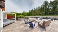 Located at 7679 Hardwood Ave S in Cottage Grove, MN, this generously sized outdoor amenity terrace offers a well-appointed communal retreat for residents of the adjoining multi-story building. The terrace is designed for both relaxation and recreation, spanning a substantial footprint across a single level.

The lounge area is anchored by a sleek concrete fire table surrounded by contemporary neutral-toned sofas and modern Adirondack-style chairs in a deep navy finish, all set atop large-format wood-look porcelain tile pavers. Additional seating groupings are arranged throughout the space, offering flexible gathering options.

A vibrant artificial turf lawn serves as the centerpiece of the recreational zone, providing a clean, low-maintenance green surface ideal for lawn games. A giant Connect Four game and a cornhole board are positioned on the turf, making the space inviting for social activity. Bold red horizontal-slat privacy screening adds a striking visual accent and defines the boundary between zones.

The terrace is enclosed by sleek black metal railings, with views extending outward to a manicured lawn area and a dense backdrop of mature trees, creating a serene, nature-connected atmosphere. The overall design balances modern aesthetics with functional outdoor living, making this terrace a standout amenity space.