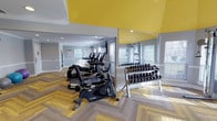 This single-floor fitness center is designed to accommodate a comprehensive workout experience, combining thoughtful architectural details with a robust selection of equipment. The space is immediately distinguished by its bold vaulted ceiling, painted in a vibrant yellow that contrasts sharply with the soft gray walls, creating an energizing atmosphere. Crown molding and a classic chair rail add a refined architectural touch throughout.

The flooring features a striking herringbone pattern in warm wood tones, lending visual interest and a polished feel to the entire space. Multiple windows fitted with white blinds allow ample natural light to fill the room, enhancing the open and airy environment.

The main exercise area is home to a row of cardio machines, including treadmills and Cybex stationary bikes, positioned for ease of use. A dedicated free weights section offers a full dumbbell rack displayed in front of a large wall mirror, allowing for form checks during training. A Life Fitness cable machine station is tucked into a well-lit corner, flanked by windows on two sides.

Additional equipment includes a rowing machine and a collection of medicine balls and stability balls along the wall, catering to functional and cross-training workouts. TRX suspension straps are also available. Recessed lighting and security cameras complete this well-appointed facility.