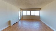 This single-floor space offers a clean, open layout highlighted by light-toned hardwood flooring that runs throughout the main living area, lending warmth and continuity to the interior. The focal point of the room is a wide bank of windows spanning nearly the full width of the exterior wall, allowing abundant natural light to fill the space and framing elevated views of the surrounding cityscape and skyline beyond. A glass-paneled door adjacent to the windows provides direct access to an outdoor balcony, further enhancing the connection between the interior and the urban environment outside. The walls are finished in a neutral beige tone that complements the flooring and contributes to an airy, understated atmosphere. A textured popcorn ceiling adds a characteristic detail to the overhead finish. A baseboard heating unit is positioned along one wall, offering a practical and unobtrusive climate control solution. The overall layout is open and unfurnished, providing flexibility for a variety of configurations. The combination of wide-format windows, elevated city views, and light interior finishes creates a bright, functional living environment well-suited to urban living.