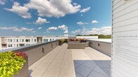 Located at 39 Trotter Road in Weymouth, Massachusetts, this rooftop deck offers a generously sized open-air retreat with a clean, modern aesthetic. The space is finished with large-format porcelain tiles that lend a polished, contemporary feel underfoot, while low-profile parapet walls define the perimeter and frame unobstructed views of the surrounding residential buildings and lush tree lines stretching into the distance.

The deck is thoughtfully arranged into distinct zones, accommodating both relaxed lounging and casual outdoor dining. Wicker and rattan furniture groupings are positioned throughout, creating comfortable gathering areas bathed in natural sunlight. Vibrant planters filled with colorful flowering plants — including reds, pinks, and greens — are distributed around the perimeter, adding lively contrast to the neutral tones of the tile and walls.

Access to the rooftop is provided through a dedicated entry door set within a white-clad exterior wall, complete with security and lighting fixtures. Utility connections and wall-mounted hardware are discreetly integrated throughout the space. The open sky above and panoramic sightlines in every direction give the deck an expansive, airy ambiance, making it an exceptional communal outdoor amenity with broad appeal for relaxation and social gatherings alike.