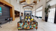 This striking clubhouse showcases an impressive architectural design anchored by soaring vaulted ceilings with exposed wood beams and black steel trusses, flooding the space with an airy, grand atmosphere. Large clerestory windows and sheer floor-to-ceiling drapery invite abundant natural light across polished large-format tile flooring. The expansive main lounge serves as the centerpiece, featuring bold contemporary furnishings including a multicolored geometric credenza, sculptural art pieces, and charcoal sofas accented with vivid yellow and teal throw pillows. A stone-clad fireplace wall with a mounted television anchors the seating area. Adjacent to the main lounge, a well-appointed library and lounge area offers floor-to-ceiling bookshelves and eclectic seating, creating an inviting retreat for reading or socializing. A dedicated game and entertainment zone features a full shuffleboard table, high-top seating, and a large-screen television set against a dramatic brushed metal accent wall. A quieter study nook provides a tufted velvet sofa, a curated gallery wall of framed artwork, and a warm wood writing desk. Entry corridors with glass double doors offer direct views to an outdoor pool area, seamlessly connecting the interior and exterior amenities. Throughout, cohesive use of teal, yellow, and neutral tones creates a dynamic yet welcoming ambiance.