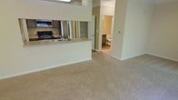 This single-floor space showcases an open-concept design that seamlessly connects the kitchen and living areas. The kitchen is equipped with black appliances, including a gas range and an overhead microwave, and features a peninsula with a built-in sink and light-toned countertops, providing both functional workspace and a natural divider between the cooking and living zones. A mirrored backsplash behind the kitchen adds depth and brightness to the space.

The living area is finished with plush, neutral-toned wall-to-wall carpeting that extends throughout, creating a warm and cohesive feel. Walls are painted in a soft green hue, contributing to a calm and inviting ambiance. Crown molding adds a subtle architectural detail overhead.

A hallway visible from the main living area leads to additional rooms, including a bathroom featuring hard flooring and standard fixtures. The layout offers a practical flow between spaces, and a thermostat is conveniently mounted on the wall near the hallway entrance. Overall, the space maintains a clean, functional aesthetic with a neutral palette suited to a variety of interior styles.