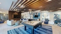 This contemporary single-story clubhouse in Gainesville, Florida, is thoughtfully designed with a cohesive blue-and-wood aesthetic that flows throughout the entire space. The expansive main lounge is anchored by striking exposed wood beam ceiling details and large floor-to-ceiling windows that flood the interior with natural light and offer views of lush surrounding greenery. Comfortable seating arrangements with modern blue upholstered chairs and accent rugs create inviting gathering areas throughout.

A central game area features a full-size billiards table set beneath a sleek pendant light fixture, adding an entertaining focal point to the space. Adjacent to the lounge is a well-appointed wet bar and entertainment area, highlighted by a dramatic floor-to-ceiling blue tile accent wall, a wall-mounted TV, and a generously sized waterfall-edge marble island with bar seating and designer pendant lighting overhead.

Private meeting rooms offer a more intimate setting, each distinguished by richly patterned blue-and-gold wallpaper, round wooden conference tables, and curated abstract artwork. The entry corridor showcases distinctive hexagonal tile flooring and a glass-walled office area, lending a polished, professional feel. A fitness area with mirrored walls and a Sputnik-style chandelier rounds out this well-appointed community facility.