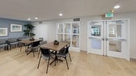 Located at 500 Harry S Truman Dr in Largo, Maryland, this single-floor community space is thoughtfully designed to accommodate group gatherings, meetings, and shared activities. The open main hall is expansive, finished with light hardwood-style flooring that creates a bright and airy atmosphere. Multiple dining and activity tables are arranged throughout, each surrounded by comfortable seating, making the space highly adaptable for a variety of uses.

The room is decorated with framed landscape artwork and lush indoor plants, adding warmth and character to the soft blue-gray walls. A wall-mounted flat-screen television, paired with an elegant wooden credenza, anchors one end of the space and serves as a natural focal point for presentations or group viewing. Rows of additional guest chairs are available nearby for organized meetings or events.

Recessed lighting and natural light from large windows ensure the interior remains well-lit and inviting throughout the day. A fully equipped kitchen area is integrated into the space, featuring stainless steel appliances including a range, microwave, and refrigerator, complemented by white cabinetry and a marble-style countertop with a sink. An accessible entry with glass partition panels connects the space to adjacent areas, supporting smooth traffic flow and a sense of openness throughout.