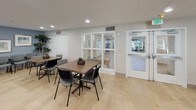 Located at 500 Harry S Truman Dr in Largo, Maryland, this single-floor community space is thoughtfully designed to accommodate group gatherings, meetings, and shared activities. The open main hall is expansive, finished with light hardwood-style flooring that creates a bright and airy atmosphere. Multiple dining and activity tables are arranged throughout, each surrounded by comfortable seating, making the space highly adaptable for a variety of uses.

The room is decorated with framed landscape artwork and lush indoor plants, adding warmth and character to the soft blue-gray walls. A wall-mounted flat-screen television, paired with an elegant wooden credenza, anchors one end of the space and serves as a natural focal point for presentations or group viewing. Rows of additional guest chairs are available nearby for organized meetings or events.

Recessed lighting and natural light from large windows ensure the interior remains well-lit and inviting throughout the day. A fully equipped kitchen area is integrated into the space, featuring stainless steel appliances including a range, microwave, and refrigerator, complemented by white cabinetry and a marble-style countertop with a sink. An accessible entry with glass partition panels connects the space to adjacent areas, supporting smooth traffic flow and a sense of openness throughout.
