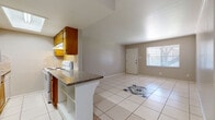 This single-floor unit offers a functional and straightforward layout designed for comfortable everyday living. The main living area is open and airy, finished with large white ceramic floor tiles that extend throughout most of the space, lending a clean and cohesive look. A decorative accent rug with a blue medallion pattern adds a touch of visual interest to the living area floor. One wall features vertical wood paneling, providing a subtle textural contrast to the otherwise neutral, smooth walls painted in soft gray tones.

The kitchen is laid out in a galley style, featuring warm-toned wooden cabinetry, a white electric range, a full-size white refrigerator, and a tile backsplash. A peninsula counter with a dark granite surface separates the kitchen from the living space, offering additional workspace and a casual breakfast bar area. A skylight overhead bathes the kitchen in natural light.

The bedroom is a private, quiet room with warm earth-toned ceramic tile flooring and a window that allows natural light to filter in. The bathroom is appointed with decorative patterned wall tiles in the shower area, a wooden vanity topped with granite countertop, a framed mirror, and neutral textured walls, completing the space with a practical yet polished finish.