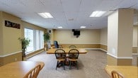 This community room is a practical and inviting shared space designed to accommodate a variety of group activities and gatherings. The room features a drop tile ceiling with recessed fluorescent lighting that ensures even, comfortable illumination throughout. Warm yellow-toned walls with a contrasting chair rail detail give the space a welcoming character, while wall-to-wall carpeting in a neutral pattern adds comfort underfoot.

At the heart of the room sits a large wooden dining-style table surrounded by spindle-back wooden chairs, providing ample seating for meetings, meals, or group activities. Additional seating is available at the periphery of the room. A wall-mounted flat-screen television serves as a focal point for presentations or shared viewing.

Large windows along one wall allow natural light to filter in, brightening the space and offering a connection to the outdoors. A decorative potted plant near the windows adds a touch of greenery. The room also includes a lounge chair tucked into a corner, offering a more relaxed seating option. Overall, the community room is thoughtfully arranged to support both structured gatherings and informal socializing in a comfortable, accessible single-floor setting.