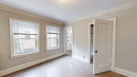 This single-floor unit showcases a thoughtful combination of classic architectural character and contemporary updates throughout. Light wood laminate flooring flows consistently across all rooms, creating a clean and cohesive aesthetic.

The main living area is bright and open, benefiting from multiple double-hung windows dressed with blinds that allow natural light to fill the space. Crown molding runs along the ceiling perimeter, lending a refined, traditional touch. A cast iron radiator adds a vintage charm while serving as a functional heating element.

The kitchen features rich, dark gray shaker-style cabinetry paired with granite countertops and a stainless steel sink. A full-size stainless steel refrigerator and additional appliances are included. An elegant arched detail and wainscoting panel on an adjacent wall add architectural interest.

The bedroom is generously sized with multiple windows providing natural light and views to the exterior. It connects directly to the bathroom, offering convenient access.

The bathroom retains its vintage character with textured walls, a classic cast iron radiator, a white vanity with a framed mirror, and subway tile surrounding the shower area. Warm brass fixtures complete the timeless aesthetic.

Overall, the space presents a warm, neutral palette with enduring architectural details that give it a distinctive and inviting character.