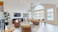 This single-floor space is thoughtfully designed with an open layout connecting several distinct functional areas. The centerpiece is a dramatic great room distinguished by soaring vaulted ceilings, a striking arched window wall that floods the space with natural light, and views of the outdoors. A globe-style chandelier anchors the room, complementing light hardwood-style flooring and neutral wall tones. Comfortable seating arranged around a round wood coffee table creates an inviting focal point, while a media console with a wall-mounted television completes the living area.

Adjacent to the great room, a casual dining nook features a round wooden table surrounded by upholstered chairs, set beneath recessed lighting and flanked by windows that continue the theme of natural light. Nearby, a kitchen bar with pendant lighting and tufted counter stools offers additional seating alongside dark cabinetry and a contrasting countertop.

A separate foyer-style area with light flooring leads to a dedicated room enclosed by classic French doors with glass panes, ideal for use as a private office or meeting space. Throughout, crown molding, recessed lighting, and cohesive neutral tones lend a polished, welcoming ambiance to the entire space.