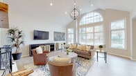 This single-floor space is thoughtfully designed with an open layout connecting several distinct functional areas. The centerpiece is a dramatic great room distinguished by soaring vaulted ceilings, a striking arched window wall that floods the space with natural light, and views of the outdoors. A globe-style chandelier anchors the room, complementing light hardwood-style flooring and neutral wall tones. Comfortable seating arranged around a round wood coffee table creates an inviting focal point, while a media console with a wall-mounted television completes the living area.

Adjacent to the great room, a casual dining nook features a round wooden table surrounded by upholstered chairs, set beneath recessed lighting and flanked by windows that continue the theme of natural light. Nearby, a kitchen bar with pendant lighting and tufted counter stools offers additional seating alongside dark cabinetry and a contrasting countertop.

A separate foyer-style area with light flooring leads to a dedicated room enclosed by classic French doors with glass panes, ideal for use as a private office or meeting space. Throughout, crown molding, recessed lighting, and cohesive neutral tones lend a polished, welcoming ambiance to the entire space.