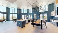 Located at 37 8th Ave in Oakland, CA, this generously sized amenity space delivers a sophisticated, resort-inspired environment across a single open floor. The design is anchored by dramatic double-height ceilings and expansive floor-to-ceiling windows that flood the interior with natural light and frame sweeping views of the surrounding cityscape and waterway.

Bold teal-blue accent walls define the space throughout, complemented by large-format neutral tile flooring that unifies the various zones. A fully equipped gourmet kitchen features stainless steel appliances including a professional-grade gas range, blue cabinetry paired with light wood upper cabinets, a glossy tile backsplash, and a built-in aquarium as a striking focal point. An adjacent prep and serving island provides ample workspace for entertaining.

The lounge areas are furnished with plush sofas, accent chairs, and coffee tables arranged around a tiled fireplace wall adorned with decorative geometric tilework and a floor-to-ceiling shelving feature. Statement pendant and disc lighting fixtures add warmth and visual interest overhead. A shuffleboard table anchors a dedicated game area, while multiple dining tables with chairs offer flexible seating. Large glass doors open to outdoor areas, enhancing the connection between interior and exterior. The overall ambiance is modern, vibrant, and highly social.