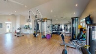 This single-floor fitness center, branded as a HealthHUB, offers a comprehensive workout environment designed to support a wide range of exercise disciplines. The space is organized into distinct functional zones, each tailored to a specific training focus.

The cardio area features a row of TRUE-brand treadmills, upright and recumbent bikes, elliptical trainers, and a stair climber. Wall-mounted flat-screen televisions are positioned throughout, and large glass doors and windows along the perimeter flood the space with natural light while providing outdoor views.

The strength and functional training zone is centrally anchored by a multi-station cable and functional training rig, complete with gymnastics rings, a rope ladder, and a heavy punching bag. Nearby, a full dumbbell rack with adjustable benches and a dedicated cable resistance machine provide additional strength training options. Exercise balls, foam rollers, and accessories are neatly stored for stretching and mobility work.

Throughout the facility, warm hardwood-style flooring, vaulted ceilings, ceiling fans, and recessed lighting create an open and inviting atmosphere. Floor-to-ceiling mirrors along one wall amplify the sense of space. A small service counter area with storage is visible, adding a practical organizational element to the overall layout.