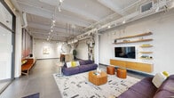 Located at 1140 E Market St in Akron, Ohio, this single-floor commercial space offers a thoughtfully designed, open-plan layout that blends industrial aesthetics with contemporary comfort. The exposed ceiling reveals structural beams, ductwork, and a track lighting system, lending the space a loft-like, urban character throughout.

The main lounge area serves as the heart of the space, anchored by plush, modular seating in deep purple and burgundy tones arranged around a sculptural wooden coffee table. A wall-mounted media console with floating wood shelving provides a polished focal point. A striking installation of large circular metal sculptural elements serves as a dramatic room divider, adding artistic depth and visual intrigue.

Large floor-to-ceiling windows along one wall flood the interior with natural light and offer views to the exterior. The entryway features glass-paneled double doors with an adjacent seating nook. A communal high-top table with leather-topped stools occupies a gallery-like area, complemented by curated wall art. A dedicated locker or changing area includes warm wood benches, storage, and a bold photographic forest mural as a dramatic backdrop. Large-format grey tile flooring ties the entire space together cohesively, creating a polished, professional ambiance throughout.
