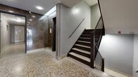 Located at 137 Isabella Street in Toronto's midtown neighbourhood, this building lobby serves as the primary point of entry for residents and visitors. The space is designed with a clean, professional aesthetic that balances functionality with refined finishes. Underfoot, speckled terrazzo flooring in warm amber and neutral tones extends throughout the corridor, lending a classic yet durable character to the space. One wall is clad in large-format, polished dark marble panels with rich veining, creating a striking architectural accent that adds a sense of elegance to the interior. The opposite walls are finished in a smooth grey tone, maintaining a contemporary and cohesive palette. Recessed ceiling lights provide consistent, even illumination along the length of the corridor, complemented by a textured white ceiling. Near the entrance, a bank of resident mailboxes is neatly integrated into the wall, offering convenient mail access. A set of dark-framed glass doors opens to the exterior, allowing natural light to filter into the entryway. An elevator provides vertical access to upper floors, while a well-maintained staircase with dark wood treads, metal balusters, and a sleek handrail offers an additional means of circulation. Security is supported by ceiling-mounted surveillance cameras positioned throughout the lobby.