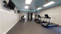 This fitness room is a dedicated, single-floor exercise space designed to support a well-rounded workout routine. The room's interior features a distinctive teal-painted ceiling paired with white beadboard wainscoting along the lower portion of the walls, creating a clean and polished aesthetic. Durable speckled rubber flooring covers the entire space, providing both safety and comfort during exercise.

The room is equipped with an array of cardio and strength equipment, including two treadmills, an elliptical trainer, a stationary upright bike, and a multi-function cable machine suitable for a variety of strength training exercises. Wall-mounted flat-screen televisions are positioned for visibility throughout the workout area, adding an entertainment option for users.

Illumination is provided by recessed rectangular LED panel lights in the ceiling, delivering bright, even lighting throughout the space. A ceiling fan contributes to air circulation and comfort during use. Natural light filters in through windows along one wall, enhancing the brightness of the room. A water cooler station is conveniently positioned near the entrance, and a full-length mirror on one wall expands the visual sense of the space while allowing users to monitor their form. The overall ambiance is functional, tidy, and well-maintained.