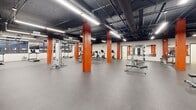 Located at 1140 E Market St in Akron, Ohio, this single-floor fitness facility offers a generously sized, open-concept layout designed to accommodate a full range of exercise activities. The main workout area is defined by bold orange structural columns that rise to an exposed black ceiling, where ductwork, piping, and industrial-style linear lighting create a modern, warehouse-inspired aesthetic. Durable rubber flooring spans the expansive main floor, providing a practical and safe surface throughout.

The cardio zone features a well-equipped row of treadmills and elliptical trainers, complemented by wall-mounted televisions and windows that bring in natural light and views of the surrounding streetscape. The strength and free weights area is lined with floor-to-ceiling mirrors, enhancing the sense of openness, and includes cable machines, weight benches, kettlebells, and resistance equipment. Ample open floor space allows for functional training and stretching.

A separate, enclosed studio room offers a distinct environment with light wood flooring, a drop tile ceiling, and bright recessed lighting — ideal for group fitness classes or personal training sessions. The facility also includes restroom access and a welcoming entry vestibule with glass doors.