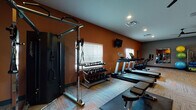 Located at 7870 Alta Dr. in Columbus, Ohio, this fitness center is a thoughtfully designed, single-floor facility built to support a wide range of workout routines. The space features a bold interior palette, with warm terracotta-orange accent walls complemented by cool gray tones, creating an energizing yet focused atmosphere. Patterned carpet tiles in neutral tones with orange accents cover the floor throughout, adding texture and visual interest while providing cushioning underfoot.

The cardio section is well-appointed with multiple Life Fitness treadmills, elliptical trainers, and stationary bikes arranged in rows, each equipped with individual display screens. A large mirror wall with floor-to-ceiling glass panels runs along one side, visually expanding the space and allowing users to monitor their form. Wall-mounted flat-screen televisions are positioned throughout for entertainment during workouts.

The strength training area includes a full dumbbell rack with a comprehensive weight range, a cable crossover machine, and a weight bench for targeted exercises. A dedicated functional training zone features stability balls, resistance bands, medicine balls, and other accessories neatly stored on a rack. Ceiling fans and recessed lighting ensure consistent comfort and visibility. The overall design balances functionality with an inviting, modern aesthetic.