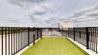 This rooftop outdoor space is designed to provide an open-air retreat above the surrounding landscape. The area is finished with lush artificial turf, offering a clean and low-maintenance green surface that stands in contrast to the concrete and metal elements of the rooftop setting. A sturdy black metal fence with a classic vertical rail design encloses the perimeter, ensuring safety while maintaining visibility to the expansive sky and surrounding rooftop views beyond. The fencing features a gated entry, allowing for controlled access to the space. Low-profile bollard-style posts are positioned along the turf boundary, further defining the area. The open-sky exposure allows for generous natural light throughout the day, making the space bright and inviting. The panoramic views of the horizon, framed by a wide, cloud-filled sky, give the area a sense of openness and scale. This rooftop area is well-suited for use as a pet relief zone, a casual outdoor lounge, or a relaxation area, combining functional design with an elevated outdoor experience above the building's immediate surroundings.