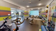 The fitness center at 103 Eudora Welty Dr in Starkville, Mississippi, is a well-equipped, single-floor facility divided into two interconnected workout zones. The space is defined by its distinctive interior design, featuring bold horizontal stripes of yellow, orange, red, and gray across the walls, complemented by crisp white crown molding and recessed lighting throughout.

The main cardio and strength area is generously sized, housing multiple treadmills, elliptical machines, and a cable/multi-use weight machine. Large mirrors line the walls, visually expanding the space and allowing users to monitor their form. Natural light enters through several windows dressed with blinds, and ceiling fans provide additional air circulation alongside the recessed fixtures above.

The second zone serves as a dedicated free weights and functional training area, equipped with a full dumbbell rack, medicine ball storage, a weight bench, and a spin bike. A large flat-screen display is mounted prominently on the wall, ideal for guided workouts or entertainment. Rubber flooring throughout both areas provides durability and safety underfoot.

An AED device is mounted on the wall, reflecting a commitment to member safety. The overall ambiance is energetic and motivating, with the vibrant color scheme and ample equipment making this fitness center a comprehensive resource for residents seeking a full range of workout options.