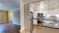 This single-floor residence showcases a thoughtfully designed interior highlighted by crown molding that adds a refined architectural touch throughout. The kitchen is a standout feature, equipped with a full suite of stainless steel appliances, including a French door refrigerator, a gas range with oven, and an over-the-range microwave. White shaker-style cabinetry extends to the ceiling, maximizing storage space, while a gray tile backsplash adds a modern contrast. Dark stone countertops provide a sleek, durable work surface. The kitchen transitions seamlessly into the adjacent living area, separated by a structural column with louvered closet doors—a functional architectural detail that also defines each space. Light hardwood flooring runs through the kitchen area, while rich, darker hardwood extends into the living space, creating a warm visual distinction between zones. Neutral warm-toned walls complement the overall palette, and flush-mount ceiling lighting ensures the kitchen remains bright and functional. The layout offers an open, connected feel while maintaining clearly defined areas for cooking and living.