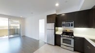 This single-floor space presents a clean, contemporary aesthetic defined by a crisp white color palette, recessed lighting, and polished flooring throughout. The kitchen is a standout feature, outfitted with a full suite of stainless steel appliances including a gas range, built-in microwave, and refrigerator. Rich, dark-toned cabinetry extends to the ceiling, providing ample storage while adding a bold contrast to the bright white walls and light countertops. A classic subway tile backsplash adds a timeless touch to the cooking area. The kitchen transitions seamlessly into an open living area, where smooth concrete-style flooring extends the modern industrial feel of the space. The open layout allows for flexible furniture arrangements, making the living area feel spacious and versatile. Natural light flows through the space, enhancing the airy, minimalist ambiance. A doorway visible from the main area leads to an additional room, suggesting a thoughtful and functional floor plan. The overall design balances modern style with practical living, making it a well-appointed and visually cohesive space.