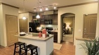 This single-floor residence is thoughtfully designed to balance style and functionality throughout its open layout. The kitchen serves as the heart of the space, showcasing rich dark cabinetry that extends to the upper walls, complemented by a classic mosaic tile backsplash and sleek granite countertops. A central island with bar seating provides an ideal spot for casual dining or entertaining, while pendant lighting and track lighting overhead create a warm, inviting atmosphere. Full-size black appliances, including a refrigerator, microwave, and range, offer a cohesive, modern aesthetic. Warm hardwood-style flooring runs throughout, tying the spaces together seamlessly. An elegant arched doorway leads from the kitchen into the bedroom area, adding a refined architectural touch that enhances the overall character of the home. The bedroom, visible through the archway, is softly lit and comfortably appointed, contributing to the cozy ambiance of the residence. Crown molding accents the ceilings, lending a polished, upscale finish to every room. The layout promotes an easy flow between spaces, making the home feel open and connected while maintaining a sense of warmth and comfort throughout.