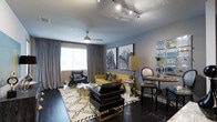 Located in Dallas, this single-floor space makes a striking impression through its carefully curated interior design. The living area showcases rich, dark hardwood flooring that grounds the room and provides a dramatic contrast to the cool grey walls and ceiling. A ceiling fan adds both function and a touch of classic detail overhead, while a sleek track lighting system illuminates the space with precision.

The furnishings strike a bold balance between glamour and comfort. A vibrant mustard yellow sofa serves as the focal point of the seating area, complemented by a tufted black leather chair and ottoman. A patterned area rug in neutral tones anchors the arrangement, while mirrored accent tables and gold-toned lamp bases introduce a refined, luxurious touch. Architectural art prints of a city skyline adorn the walls, nodding to the urban Dallas setting.

Flowing grey drapery panels frame a bright window, softening the space and allowing natural light to filter through. A mirrored bar display unit in the corner offers an elegant vignette for glassware and barware. A small dining or bar-height table with bistro-style chairs rounds out the space, making it as functional as it is visually dynamic. The overall ambiance is sophisticated, bold, and unmistakably urban.