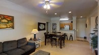 This single-floor residence offers a well-designed open-concept layout that seamlessly connects the living, dining, and kitchen areas into one cohesive and functional space. The living area is warmly appointed with a plush sofa, an accent side table with a table lamp, and a ceiling fan with integrated lighting that provides both comfort and illumination. A decorative framed mirror adds a stylistic touch to the neutral-toned walls, while soft carpeting underfoot enhances the room's cozy ambiance. The dining area sits centrally within the open plan, featuring a dark wood dining table paired with matching bar-height chairs, creating a welcoming spot for meals and gatherings. A built-in bookshelf nearby offers convenient storage and display space. The kitchen, visible from the main living area, is equipped with medium-toned wood cabinetry, a white refrigerator, and a light-colored countertop that extends into a raised breakfast bar with bar stool seating — ideal for casual dining or conversation. Recessed and overhead lighting in the kitchen ensures a bright and practical workspace. The overall color palette of warm beiges and neutrals, combined with the open layout and thoughtful furnishings, creates a comfortable and inviting atmosphere throughout the space.
