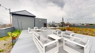 Located at 1221 N Fourth St in Philadelphia's vibrant 19122 neighborhood, this rooftop terrace offers an exceptional outdoor retreat above the city. The space is divided into two distinct zones: a generous composite deck area and an expansive living green roof planted with a colorful tapestry of sedum and low-growing succulents in shades of gold, red, and green, adding both visual interest and environmental benefit.

The deck area is thoughtfully furnished with multiple groupings of modern white-framed outdoor sofas and lounge chairs with plush gray cushions, providing ample seating for gatherings of various sizes. Overhead, strands of café lights are strung between poles, creating an inviting ambiance for evening use. The perimeter is enclosed by a clean gray panel surround that offers privacy while maintaining an open, airy feel.

From the rooftop, panoramic views extend across the Philadelphia cityscape, with the downtown skyline and its landmarks — including a notable church steeple — clearly visible. The surrounding neighborhood rooftops and tree canopy further enrich the urban backdrop. A rooftop access structure with a metal roof anchors one corner of the space. This rooftop terrace is a standout amenity, combining sustainable green design with comfortable, well-appointed outdoor living in the heart of Philadelphia.