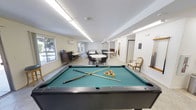This single-floor recreational space is thoughtfully designed to serve as a social hub for games, gatherings, and relaxation. At the forefront of the room stands a full-sized billiards table with a green felt surface, complete with cues and racked balls, making it a natural centerpiece for the space. Overhead, long linear fluorescent light fixtures illuminate the room evenly, complementing the natural light that flows in through a row of large windows along one wall, offering views of the surrounding greenery outside. The walls are finished in a clean, neutral tone, contributing to the bright and open atmosphere. Toward the back of the room, multiple round tables with chairs are arranged to accommodate group seating, ideal for casual dining, card games, or community events. A wall-mounted air conditioning unit ensures year-round comfort. Additional furnishings include a wooden desk area with a monitor, a cue rack mounted on the wall, and a cushioned accent chair tucked neatly in the corner. A corridor leads further into the facility, suggesting connectivity to additional amenities. Overall, the space strikes a balance between functionality and comfort, making it well-suited for a variety of recreational and social activities.