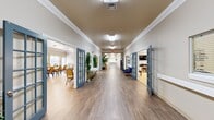 This expansive single-story care community facility offers a welcoming and well-appointed environment designed for comfort and engagement. The layout is anchored by a broad central corridor finished with light hardwood-style flooring, crown molding, and distinctive blue French doors that open into various activity and common spaces. Decorative white columns add an elegant architectural touch throughout.

The facility includes a generously sized dining and activity hall with ample seating, multiple tables, and bright overhead lighting — ideal for group gatherings and community events. An adjacent serving area features a whiteboard, beverage stations, and prep counters to support daily programming. A cozy lounge area offers carpeted flooring, wall-mounted television, framed artwork, and comfortable seating for relaxed socializing.

A dedicated activity room provides round tables with vibrant teal and white chairs, shelving with games and resources, and a cheerful décor. A library and multi-purpose room houses bookshelves, craft storage, and a podium, reflecting diverse programming options. A full-service salon with styling equipment and nail care stations adds a personal wellness amenity. Private meeting rooms with carpeted floors, crown molding, and tasteful décor round out the offerings, creating a comprehensive and thoughtfully designed community space.