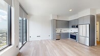 Located in Fort Lauderdale's vibrant urban core, this contemporary studio offers a thoughtfully designed open-plan layout with a clean, modern aesthetic throughout. Light hardwood flooring runs seamlessly across the entire space, complementing the crisp white walls and creating a bright, airy atmosphere.

The living area is anchored by expansive floor-to-ceiling windows fitted with roller shades, flooding the space with natural light and framing striking panoramic views of the Fort Lauderdale skyline. A distinctive exposed concrete structural column adds an industrial character to the otherwise minimalist interior.

The kitchen is well-appointed with flat-front gray cabinetry extending to the ceiling for ample storage, a white countertop, and a full suite of stainless steel appliances including a gas range, over-the-range microwave, dishwasher, and French door refrigerator. A matte black faucet and undermount sink complete the modern kitchen design.

The bathroom features large-format stone-look wall tiles, a mosaic tile shower floor with a center drain, a glass-enclosed walk-in shower, and a wall-mounted toilet, all contributing to a refined, spa-inspired feel. A dedicated closet provides convenient storage. This space is ideal for those seeking a stylish, low-maintenance urban residence in Fort Lauderdale.