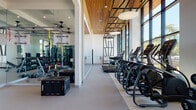 This single-floor fitness center in Portland, Oregon, is designed with both functionality and aesthetics in mind. The space is defined by soaring floor-to-ceiling glass windows along its exterior wall, flooding the interior with abundant natural light and providing open views of the surrounding outdoor landscape. The architectural design incorporates a striking wood-slat ceiling that adds warmth and texture, complementing the clean white walls and smooth neutral-toned flooring throughout.

The cardio zone is well-equipped with a row of treadmills, elliptical trainers, stair climbers, upright cycling bikes, and a recumbent bike, all positioned to take full advantage of the expansive window views. A dedicated strength and functional training area features floor-to-ceiling mirrors, TRX suspension systems, a cable machine, a rack of dumbbells, weight benches, and plyo boxes, offering a complete training environment. A decorative geometric black-framed wall accent serves as a visual focal point between zones.

Large pendant light fixtures hang from the wood ceiling, providing even illumination alongside recessed lighting. The overall ambiance is bright, modern, and well-organized, creating an inviting and professional environment suited for a full range of fitness activities.