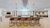 This thoughtfully designed single-floor amenity space offers a diverse range of social and recreational areas unified by a clean, contemporary aesthetic. Entry is made through striking floor-to-ceiling black-framed glass double doors that immediately reveal the heart of the space: a generous central bar island topped with sleek white quartz countertops and surrounded by modern red and tan barstools. Pendant lighting in mixed gold and glass finishes hangs above, complemented by an eye-catching wood-paneled ceiling detail that adds warmth and architectural character. Structural columns with wood accents frame the open layout, while large windows allow abundant natural light to fill the room. A full-length shuffleboard table and a wall-mounted flat-screen TV anchor the game and entertainment zone, creating an inviting hub for social activity. Adjacent to the main area, a cozy lounge nook features soft seating and curated artwork in a more intimate, subdued setting. A dining area with upholstered booth seating and pendant lighting provides a casual yet refined space for group gatherings. A secondary lounge room, finished in warm taupe tones with light wood flooring, offers a quieter retreat. Throughout, light-toned flooring, recessed lighting, and a neutral color palette create a cohesive, welcoming ambiance suited for community living.