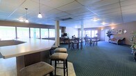 This single-floor space is a generously sized gathering and dining area designed to accommodate groups comfortably. The room is anchored by a prominent bar counter with high bar stools along one side, making it well-suited for casual socializing or events. Throughout the open floor plan, multiple high-top tables paired with bar-height chairs are arranged to maximize seating capacity while maintaining an open, airy feel. The patterned green carpet adds a classic touch to the interior and helps define the space underfoot. A drop ceiling with recessed grid tiles and a combination of pendant and flush-mount lighting provides consistent, even illumination throughout. Large windows line the perimeter walls, allowing abundant natural light to pour into the space and creating a bright, welcoming atmosphere. Structural columns are thoughtfully integrated into the layout without disrupting the flow of the room. A media station or service counter is visible toward the back of the space, adding a functional element for events or presentations. Overall, the layout is flexible and inviting, suitable for dining, receptions, or community gatherings.
