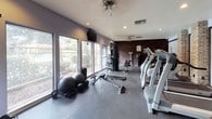 This fitness center offers a comprehensive workout environment designed to accommodate a wide range of exercise routines. The space is defined by a distinctive combination of exposed brick walls and dark wood-paneled diagonal accent walls, lending the facility a warm, classic character that sets it apart from a typical gym setting. Rubber flooring throughout provides a durable, functional surface suited for both strength and cardio training.

A full dumbbell rack lines the brick wall near the entrance, offering an organized selection of free weights for resistance training. Adjacent to it, a cable machine with rope attachments provides versatile upper-body workout options. The main workout floor features multiple cardio machines, including ellipticals and a stair climber, as well as an adjustable weight bench and stability balls for functional fitness. A multi-station cable and weight machine anchors the center of the room, supporting a variety of strength exercises.

Large windows along one wall flood the space with natural light and offer views of the surrounding greenery, creating an open and refreshing atmosphere. Ceiling fans and recessed lighting ensure comfort and visibility throughout. A classic arched glass door at the entrance connects the facility to an exterior walkway, completing this well-appointed and inviting fitness space.