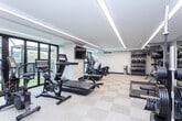 Located at 185 N West Temple in Salt Lake City, UT, this well-appointed fitness center offers a spacious, single-level layout designed to support a full range of workout routines. The exercise room is finished with a neutral-toned carpet tile floor and features a clean, modern aesthetic throughout.

Linear LED lighting runs across the ceiling in sleek parallel strips, providing bright, even illumination across the entire space. Large floor-to-ceiling windows allow ample natural light to filter in while offering views of the outdoor greenery, creating an energizing atmosphere. Full-length mirrors line one wall, enhancing the sense of openness and allowing users to monitor their form during workouts.

The facility is comprehensively equipped with a variety of Freemotion and Technogym strength machines, including a functional trainer, a cable system, and a leg press. A dedicated free weights area features a full rack of dumbbells and a barbell station with weight plates. Cardio options include treadmills, stationary bikes, and a rowing machine. Wall-mounted flat-screen TVs provide entertainment throughout the space. A built-in wooden shelving unit offers convenient storage for accessories, and a water fountain station is available for hydration. Direct access to an adjacent outdoor area further enhances the fitness experience.