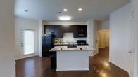 This single-floor space is designed with an open-concept layout that seamlessly integrates the kitchen and living areas. The kitchen serves as a focal point, anchored by a large central island topped with light granite countertops that provide ample prep and gathering space. Rich, dark espresso cabinetry lines the walls, extending to the upper portion of the kitchen and offering generous storage. Black appliances, including a refrigerator, range, and built-in microwave, complement the cabinetry for a cohesive, modern aesthetic. Recessed lighting and a flush-mount ceiling fixture provide bright, even illumination throughout the kitchen area. The flooring throughout the space consists of dark hardwood, adding warmth and continuity. A window along one wall allows natural light to filter into the space. An open passage leads to an adjacent room with a paneled door, suggesting additional living or bedroom space beyond. The overall design balances functionality and style, with a clean, contemporary finish palette of dark cabinetry, neutral walls, and contrasting light countertops creating a polished, well-appointed interior.