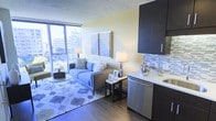 This single-floor unit showcases a thoughtfully designed open-concept layout that seamlessly integrates the living and kitchen areas. The living space is anchored by a large floor-to-ceiling window that floods the room with natural light while framing expansive city skyline views, creating a strong connection to the urban environment outside. The hardwood-style flooring runs throughout, adding warmth and continuity to the space.

The kitchen is positioned along one wall and features dark espresso cabinetry that extends to the ceiling, offering ample storage. A decorative mosaic tile backsplash adds texture and visual interest, complementing the stainless steel appliances, including a dishwasher. The countertop integrates a sink, completing this efficient and stylish cooking space.

The living area accommodates a blue upholstered sofa, side tables, and a geometric patterned area rug, all arranged to take advantage of the natural light and city views. Framed artwork on the accent wall adds a curated aesthetic. The overall ambiance is modern and cosmopolitan, with a neutral color palette punctuated by bold dark accents.