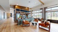 Located at 2925 Tutt Blvd in Colorado Springs, Colorado, this single-floor community amenity space offers a thoughtfully designed interior that blends modern comfort with warm, natural materials. Wide-plank light oak flooring runs throughout, complemented by exposed wood beam accents and rich reclaimed wood wall paneling that lend the space a contemporary yet inviting character.

At the heart of the space sits a prominent market-style café bar finished in sage green, featuring open shelving, a beverage cooler, and a curated display of snacks and goods. Adjacent lounge seating—anchored by cognac leather armchairs and a sculptural low coffee table—creates an informal gathering area ideal for socializing or relaxing.

Floor-to-ceiling steel-framed windows and glass partitions line key areas of the space, flooding the interior with abundant natural light and framing views of the surrounding outdoor courtyard and community. A dedicated dining area features dark marble-topped tables paired with wood and leather chairs beneath a statement chandelier, while a quieter nook with plaid built-in banquette seating offers a more intimate setting.

A co-working corridor, stylish entry benches with patterned upholstery, and carefully selected artwork round out this well-appointed, multifunctional community hub.