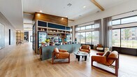 Located at 2925 Tutt Blvd in Colorado Springs, Colorado, this single-floor community amenity space offers a thoughtfully designed interior that blends modern comfort with warm, natural materials. Wide-plank light oak flooring runs throughout, complemented by exposed wood beam accents and rich reclaimed wood wall paneling that lend the space a contemporary yet inviting character.

At the heart of the space sits a prominent market-style café bar finished in sage green, featuring open shelving, a beverage cooler, and a curated display of snacks and goods. Adjacent lounge seating—anchored by cognac leather armchairs and a sculptural low coffee table—creates an informal gathering area ideal for socializing or relaxing.

Floor-to-ceiling steel-framed windows and glass partitions line key areas of the space, flooding the interior with abundant natural light and framing views of the surrounding outdoor courtyard and community. A dedicated dining area features dark marble-topped tables paired with wood and leather chairs beneath a statement chandelier, while a quieter nook with plaid built-in banquette seating offers a more intimate setting.

A co-working corridor, stylish entry benches with patterned upholstery, and carefully selected artwork round out this well-appointed, multifunctional community hub.